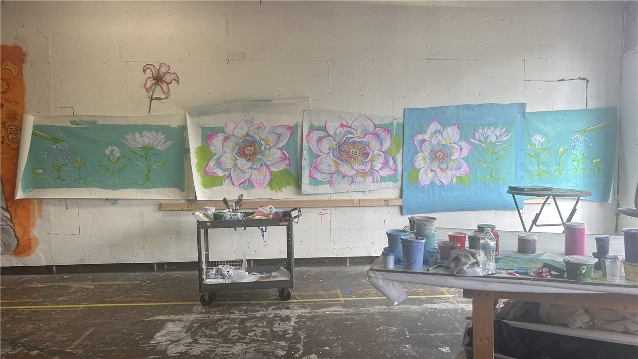 Five mural panels pinned along the studio wall in progress, lotus flowers painted on teal and blue grounds, paint carts and pots in the foreground.