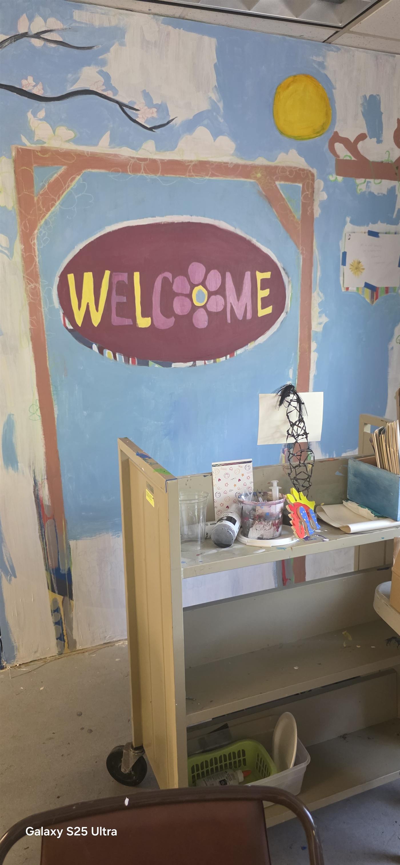 Inside the DAMA studio: a hand-painted WELCOME sign on a blue sky mural above a supply cart.