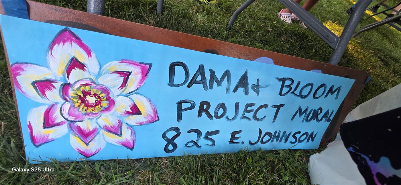 A hand-painted sign reading DAMA + Bloom Project Mural, 825 E Johnson, with a lotus flower painted beside the lettering.