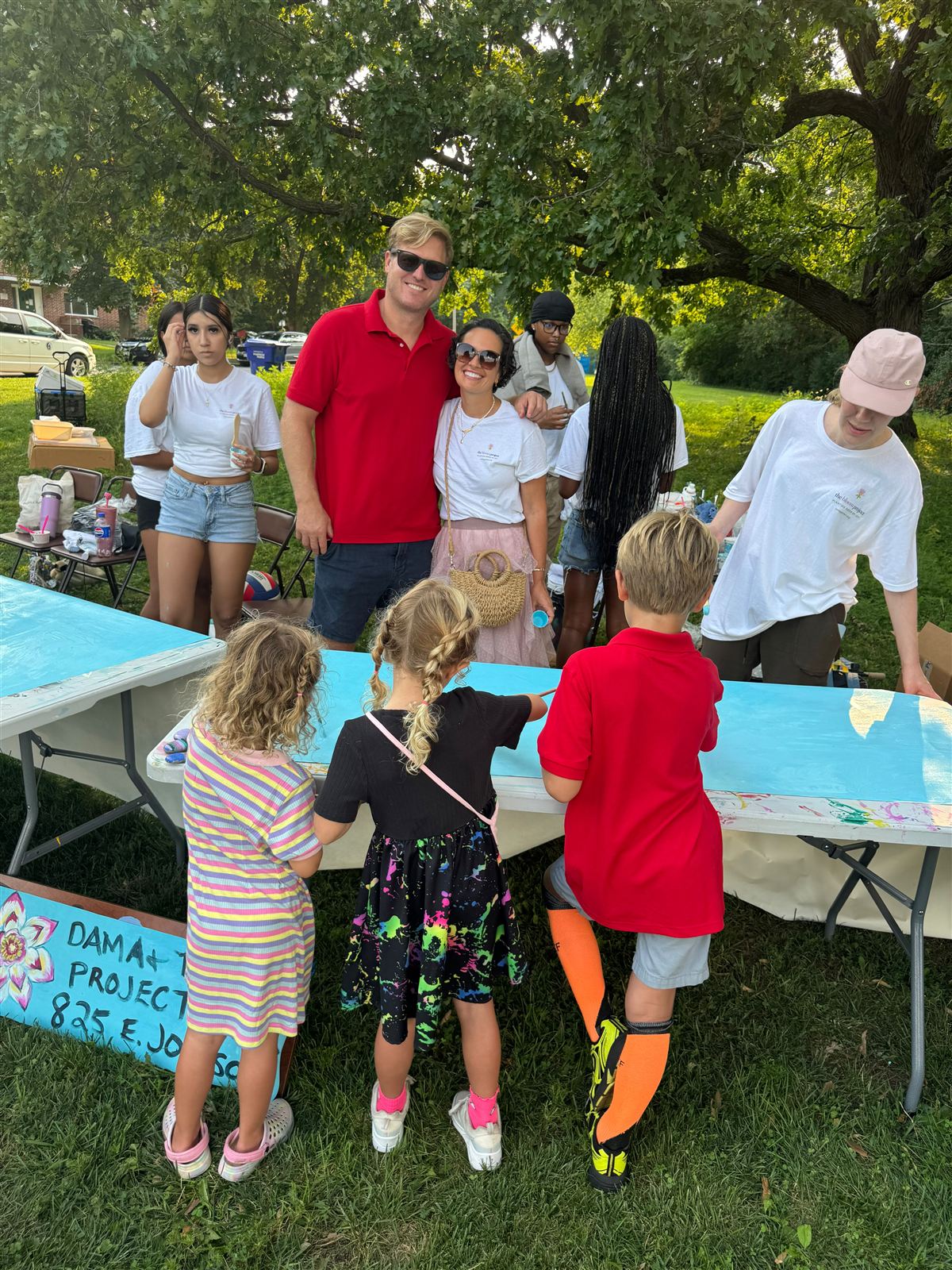 A large group gathered around the painting tables under trees at Warner Park, the hand-painted project sign propped at the end.
