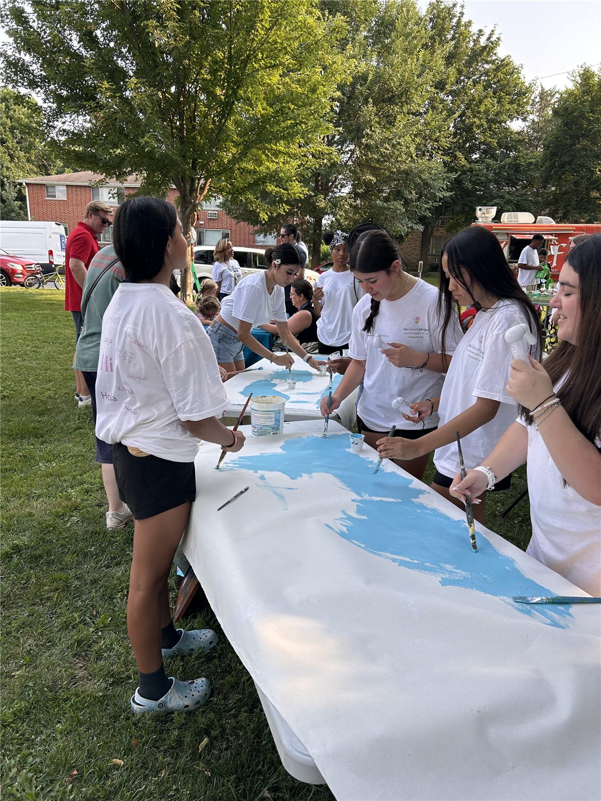 A row of interns in the multilingual project shirts leaning over a panel, laying blue paint across it.