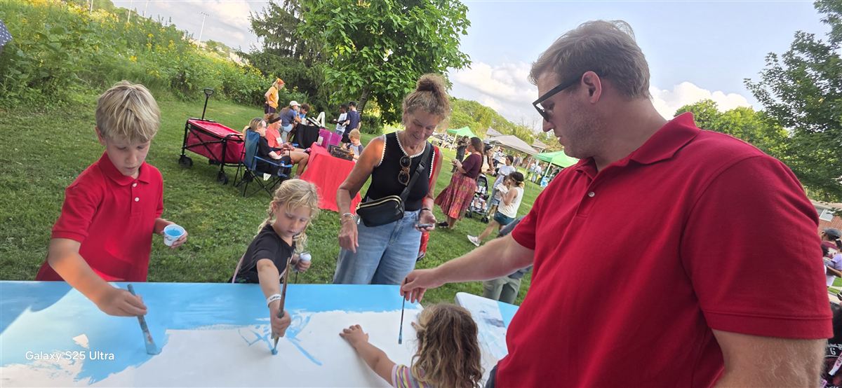Children reaching across a panel with brushes while adults steady it, a park full of people behind them.