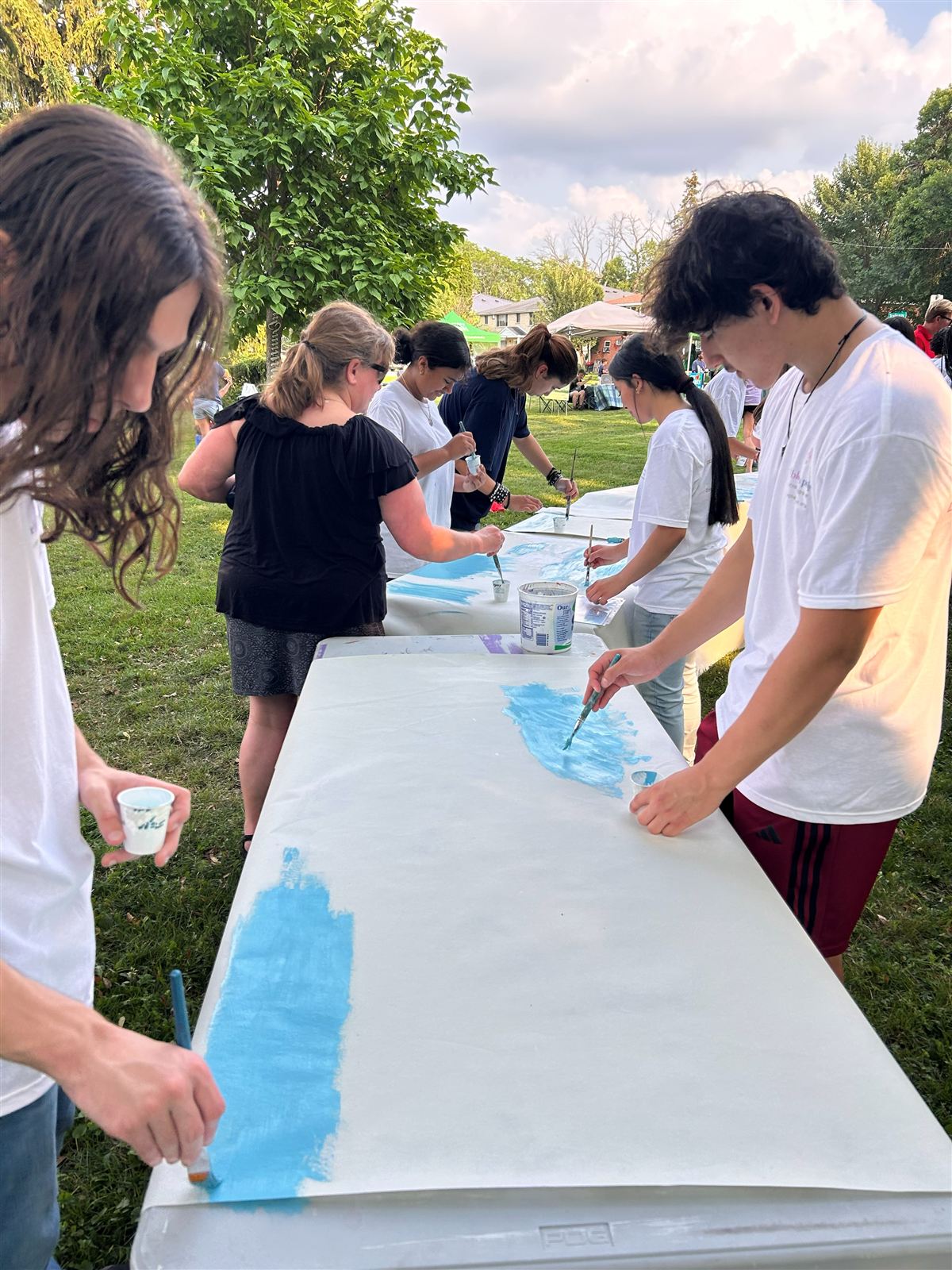 People of all ages leaning over a long panel on trestle tables in a park, brushing blue paint across it.