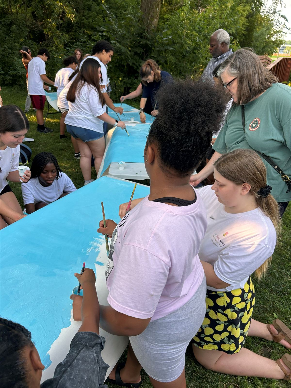 A crowd of young people and adults around two long tables, painting the blue background of the mural panels.