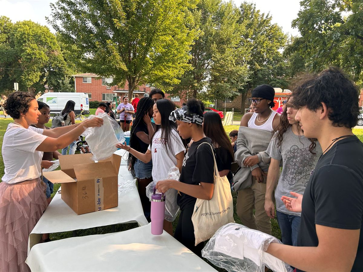 Project shirts being handed out from a cardboard box to a line of young people at the park.